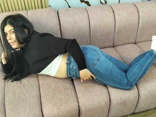 AnaTaylor est allongée sur un canapé rose, vêtue d'un pull noir, d'un haut blanc et d'un jean bleu, avec les fesses en l'air et les mains derrière le dos, regardant sur le côté..