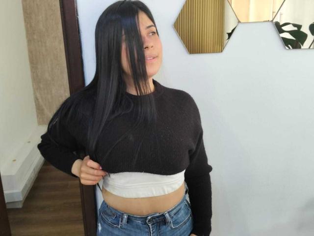 Ana Taylor mit langen, schwarzen Haaren, die einen schwarzen Pullover und eine weiße Unterlage trägt, hebt den Pullover an, um ihren nackten Bauch und blaue Jeans zu zeigen. Ihre Hand ist in ihrem Haar..