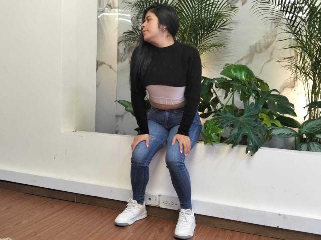 AnaTaylor, portant un haut noir à manches longues, un soutien-gorge rose, un jean bleu déchiré et des baskets blanches, assise les mains sur les genoux, regardant sur le côté, sur un plancher en bois brun..