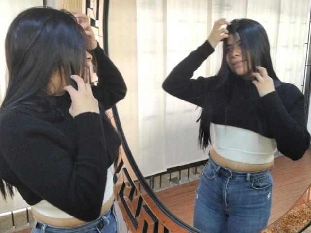 Ana Taylor mit langem, schwarzem Haar, einem schwarzen Pullover, einem weißen Crop-Top und blauen Jeans, die sich im Spiegel betrachtet und mit einer Hand ihr Haar berührt..