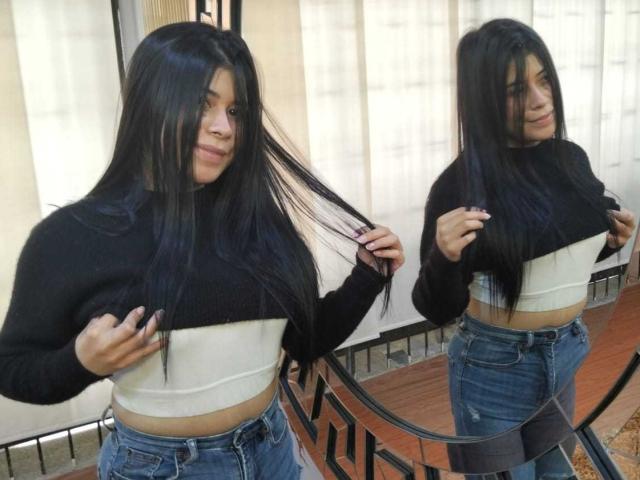 AnaTaylor with long black hair, lifting her black and white crop top, showing her small tits, wearing blue jeans, reflected in a mirror. Her hand's in her hair.