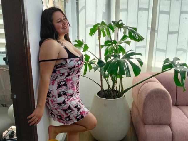 EmilyColeman69, with black hair, wearing a pink and white patterned dress, black bra, leaning against wall, smiling, barefoot, standing next to potted plant and pink sofa.