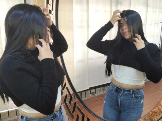 AnaTaylor with long black hair, black sweater, white crop top, and blue jeans, stands in front of mirror, touching her hair, reflection shows her smiling, her ass slightly visible.