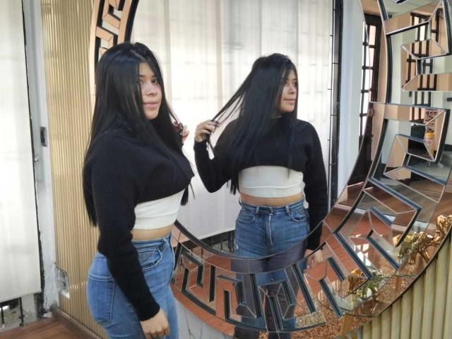 Ana Taylor, con cabello largo y negro, vestida con una blusa negra, un top corto blanco y jeans azules, está de pie frente a un espejo, sosteniendo su cabello, y se refleja su trasero..