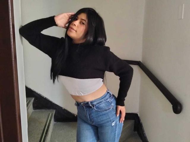 AnaTaylor mit langen, schwarzen Haaren, einem schwarz-weißen Crop-Top und blauen Jeans, steht auf einer Treppe, eine Hand am Kopf, blickt zur Seite und zeigt einen schlanken, hübschen Hintern..