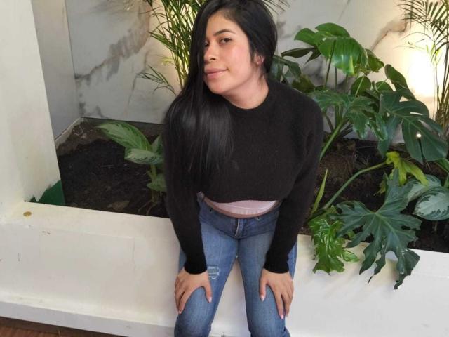 AnaTaylor mit langem, schwarzem Haar, einem schwarzen Pullover und blauen Jeans, sitzt, die Hände auf den Knien, blickt zur Seite, trägt einen kleinen, pinken Bund, im Hintergrund sind grüne Pflanzen zu sehen, der Boden ist aus Holz..