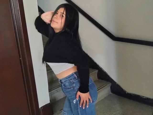AnaTaylor mit langen, schwarzen Haaren, einer schwarzen, kurzen Kapuzenjacke, einem weißen Oberteil und blauen, zerrissenen Jeans, steht auf einer Treppe, eine Hand auf dem Hintern, blickt über die Schulter, lächelt und trägt ein schwarzes Armband..
