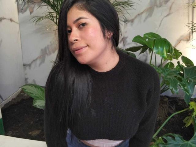 AnaTaylor, avec ses longs cheveux noirs, son pull noir et sa ceinture rose, souriant légèrement, se tient près de plantes vertes, montrant ses fesses et ses petites seins, dans un cadre intérieur décontracté..