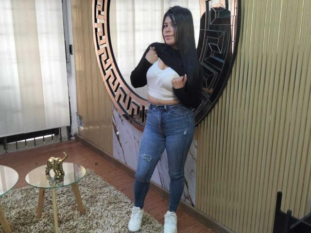 AnaTaylor, avec ses longs cheveux noirs, soulève un t-shirt noir pour montrer son crop top blanc, son jean bleu déchiré et ses baskets blanches, et se tient dans une pièce avec un mur doré..