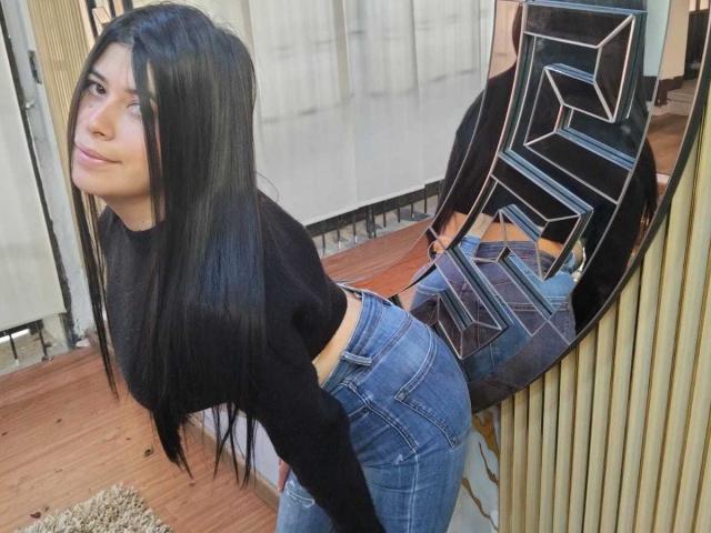 AnaTaylor met lang zwart haar, een zwarte crop top en blauwe jeans, gebogen, waarbij haar achterwerk zichtbaar is, voor een spiegelwand die haar reflecteert..