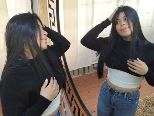 Ana Taylor, con cabello negro largo, vestida con una blusa negra y un top corto blanco, jeans azules, con una mano en la cabeza y otra en el pecho, sonriendo, reflejo en un espejo, piso de madera, atuendo casual..