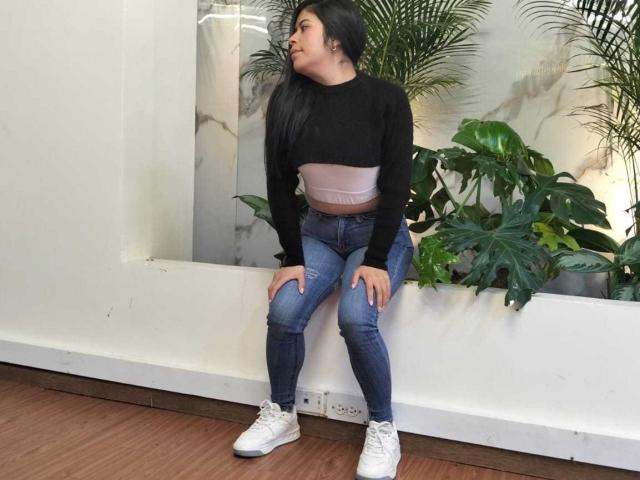 AnaTaylor, black long-sleeve top, pink ribbed crop, blue jeans, white sneakers, sitting, hands on knees, looking to the side, brown wooden floor, green plants, white wall.