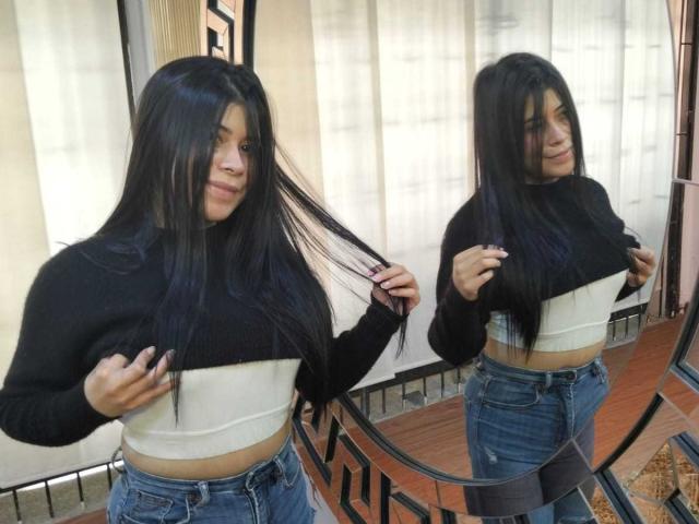 AnaTaylor, con lunghi capelli neri, indossa una maglietta nera e un top corto bianco, jeans blu, tiene i capelli e si trova di fronte a uno specchio, mostrando il suo riflesso..