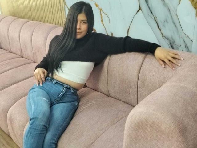 AnaTaylor, avec ses longs cheveux noirs, portant un pull noir court, un débardeur blanc et un jean bleu, est assise sur un canapé rose, sa main posée à l'arrière..