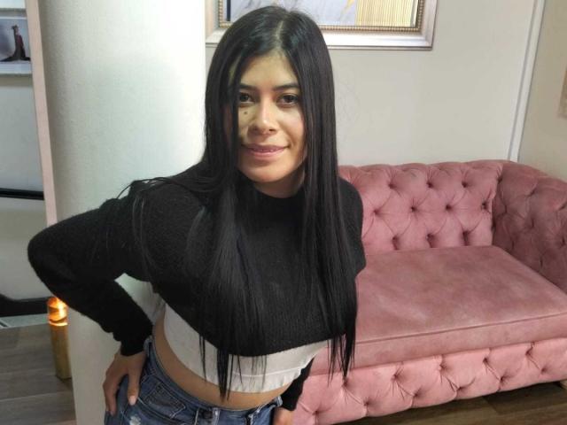 AnaTaylor with long black hair, wearing a black crop top and blue jeans, standing with hands on hips, smiling slightly, pink tufted couch in background.