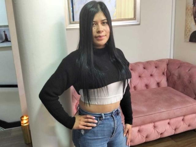 AnaTaylor mit langen, schwarzen Haaren, die ein schwarzes Crop-Top und blaue Jeans trägt, steht vor einem rosa, gepolsterten Sofa, die Hände in die Hüften gestemmt und lächelt leicht..