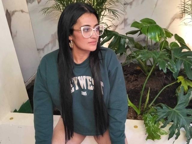 BriaVolkova, with long black hair, glasses, and green sweatshirt, sits with bare thighs, wearing gold earrings, and a slight smile. She has a cute, sexy look.