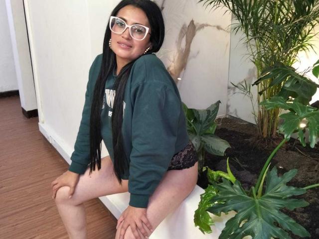 BriaVolkova with long black hair, white glasses, green hoodie, black lace panties, sitting on white bench, smiling, showing her ass, next to potted plants, on wooden floor.