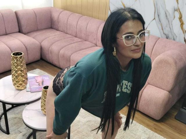 BriaVolkova with long black hair, glasses, green sweatshirt, and black lace panties, leaning forward, showing her ass, in a room with pink couch and gold-patterned vase.