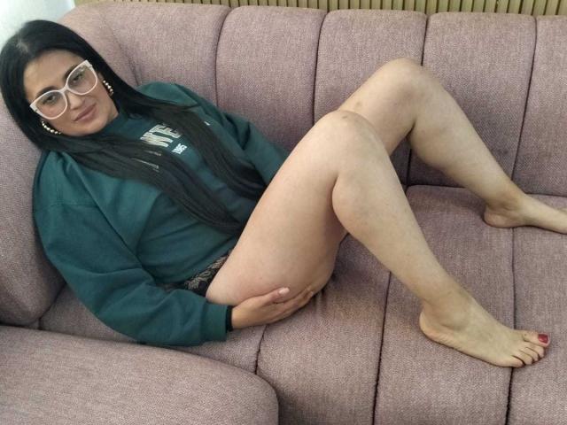 BriaVolkova, with long black hair, wearing glasses and a green hoodie, sits on a pink sofa, legs spread, showing her bare ass and red-painted toenails.