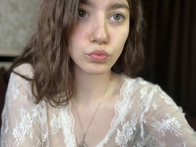 VinnaSkyHott with pouty pink lips, wavy brown hair, and light skin, wearing a sheer white lace top, showing small tits, and a silver necklace.