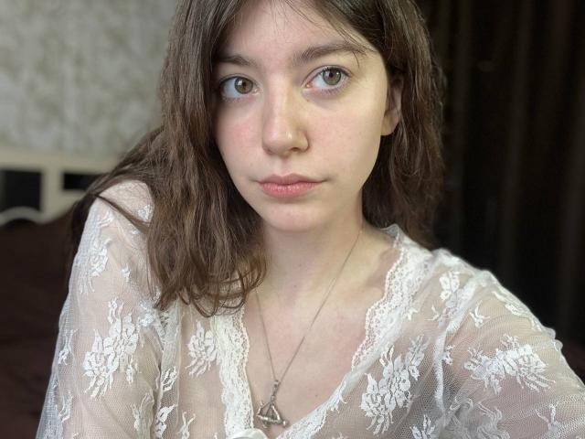 VinnaSkyHott with brown hair, green eyes, and pinkish skin, wearing a white lace top, showing small tits, and a silver necklace with a triangular pendant.