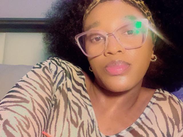 BootyBangerGoldie mit lockigem Haar, die eine Brille und ein zebra-gemustertes Hemd trägt, hat rosafarbene Lippen und einen grünen Lichtreflex in ihrem rechten Auge, sowie ein leopardenmuster-Haarband..