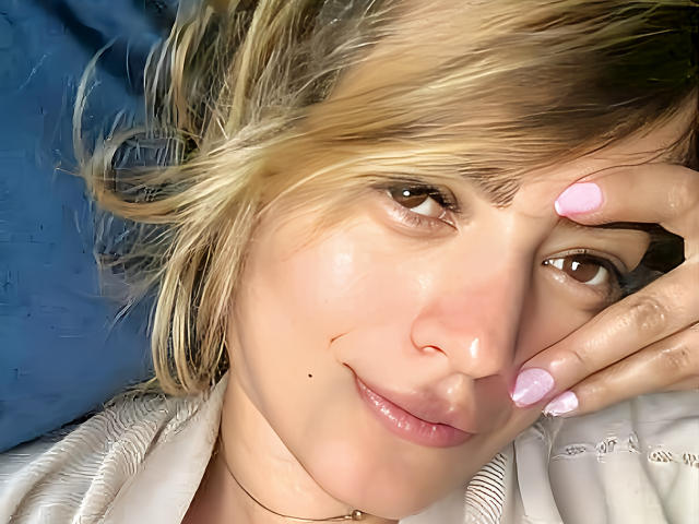 KristenSexy has blonde hair, pink nails, and a small mole on her cheek. She's smiling with her hand on her face, wearing a white shirt. Her tits are not visible.