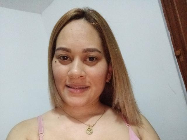 Georgie tiene el cabello castaño claro, una pequeña marca de nacimiento debajo del ojo derecho y un sujetador rosa. Está sonriendo y lleva un collar dorado con un pequeño dije circular..