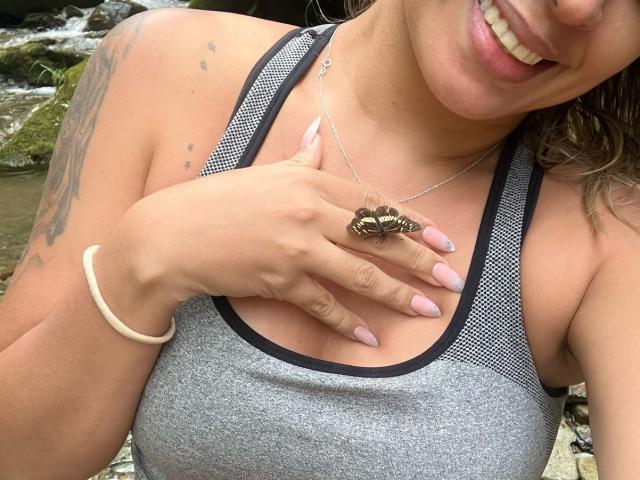 ChantalSex69 with tan skin, smiling, wearing a grey sports tank, hand on chest, showing a butterfly ring, long pink nails, white bracelet, and tattoo on right arm.