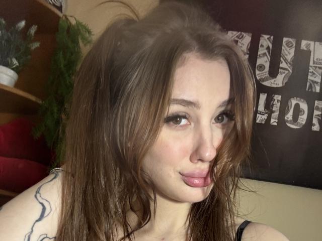 SensualMila with wet brown hair, pouty pink lips, and a tattoo on her arm, looking directly at the camera. Her tits are small and she has a cute face.