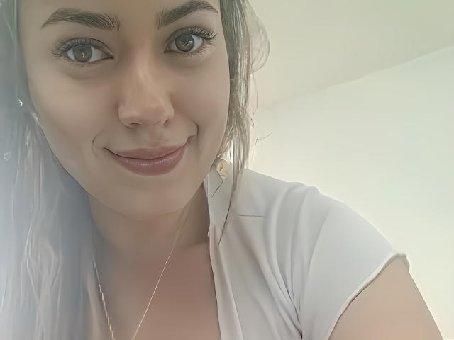 KristenSexy with light skin, grey eyes, and a slight smile, wearing a white shirt, has a small nose and is looking at the camera with a subtle smirk.