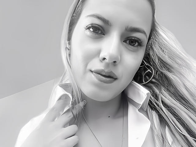 Black-and-white selfie of KristenSexy with long, straight hair, wearing a slightly open shirt, holding her collar, showing a small mole on her neck, and looking at the camera.