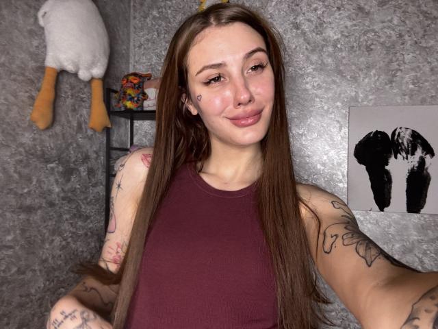 SensualMila, with long brown hair, wearing a maroon sleeveless top, has tattoos on arms and face, smiling slightly, showing her nose ring and small tattoos near her lips.
