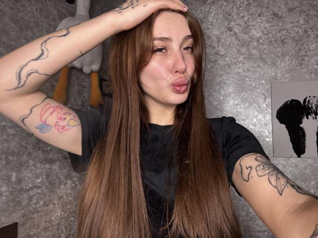 SensualMila mit langen, braunen Haaren, die die Lippen leicht zusammenpresst, trägt ein schwarzes Hemd, hat Tattoos an den Armen, eine Hand am Kopf, braune Augen, einen weichen Gesichtsausdruck, im Hintergrund eine graue, strukturierte Wand..