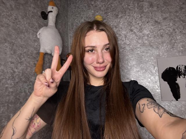 SensualMila with long brown hair, black shirt, tattoos on arms, smiling, pink blush on cheeks, making a peace sign, standing in front of a gray textured wall.