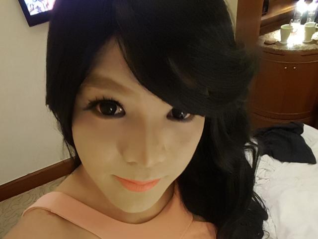 QueenOfPainSasha with black wavy hair, pale skin, and pink lipstick, wearing a pink top, showing her face and upper chest, looking at the camera with a slight smile.