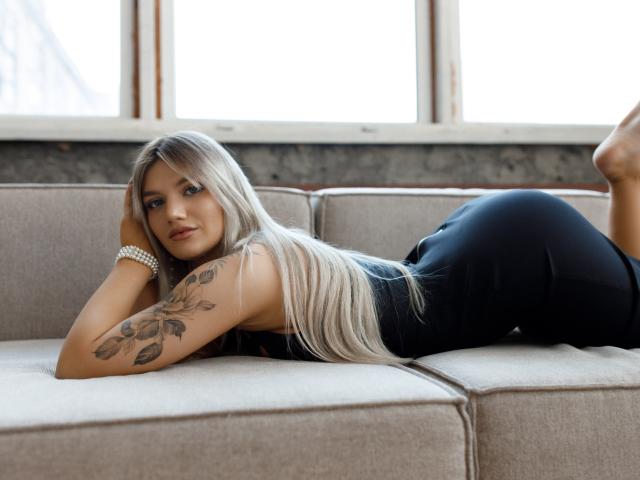 SelenaeMoore, laying on a beige couch, in black pants, with long blonde hair, tattooed arm, and pearl bracelet, looking at camera, ass raised, bare feet.