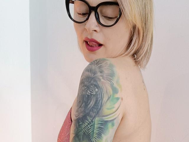 KatParadise, avec ses cheveux blonds, ses lunettes noires et son rouge à lèvres rouge, présente un tatouage détaillé d'un Ewok noir et poilu avec des feuilles vertes sur son épaule, regardant par-dessus son épaule..