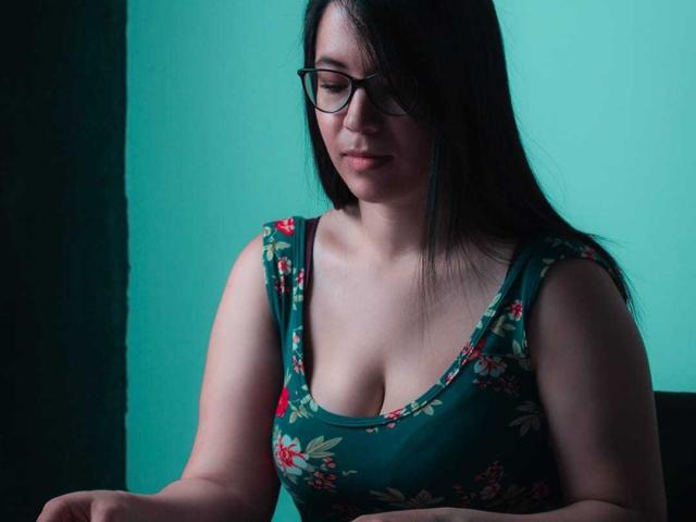 LizBellaa with glasses, dark hair, wearing a green floral tank top, showing her tits, looking down with a slight smile, in a dimly lit room.