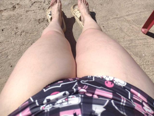 LizBellaa's pale, chubby thighs in pink and black plaid swim shorts, wearing beige flip-flops, standing on sandy beach, with a small red object in the top right corner.