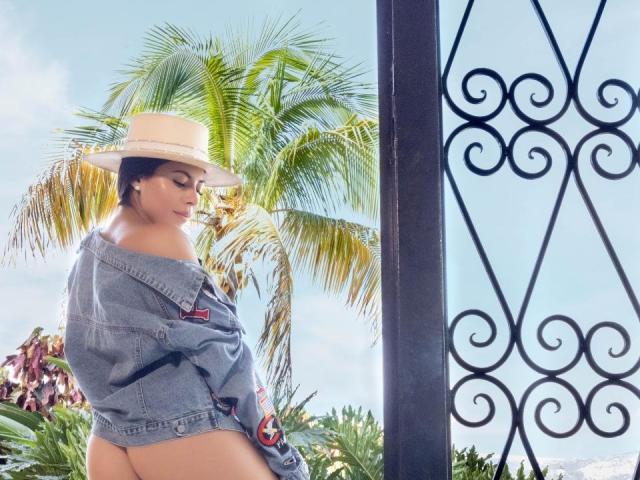 Mujeres con piel morena, senos pequeños y glúteos redondeados, vestidas con una chaqueta vaquera llevada de lado y un sombrero blanco, de pie frente a una puerta de hierro negro y una palmera..