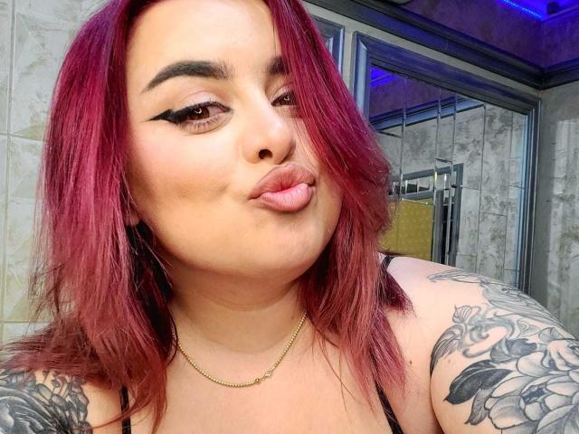 ChaudePourxToi mit roten Haaren, voller Lippen, schwarzem Eyeliner, Tattoos an den Armen, trägt ein schwarzes Oberteil, eine goldene Halskette und schaut in die Kamera. Sie befindet sich in einem Badezimmer mit einer Glasdusche..