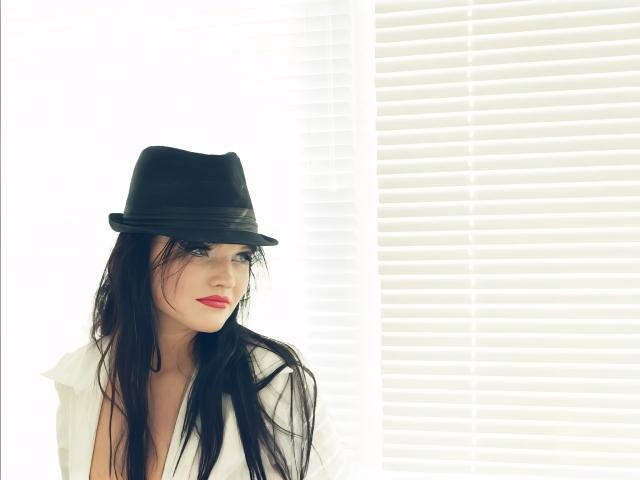 ElenaXRus, con cabello negro largo, lápiz labial rojo y un sombrero negro, vestida con una camisa blanca, mirando hacia un lado con una expresión ligeramente triste. Persianas de ventana brillantes en el fondo..