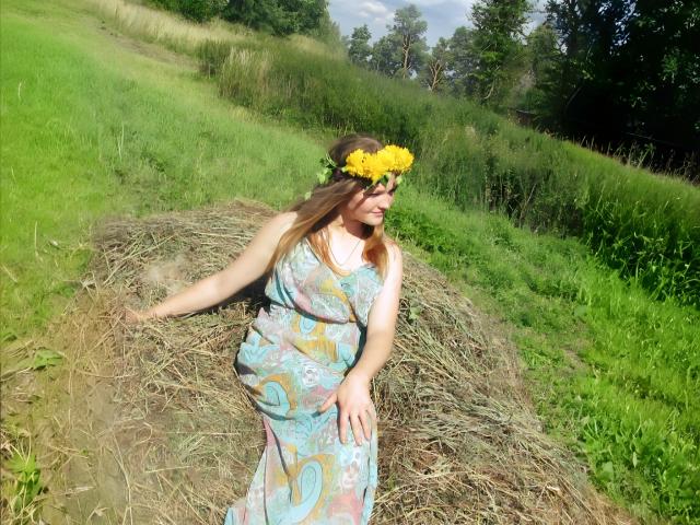 ElenaXRus, avec une couronne de fleurs jaunes, assise sur une botte de foin, portant une robe à motifs bleus et roses, sur une colline herbeuse, ensoleillé, herbe verte, arbres en arrière-plan, regardant sur le côté..