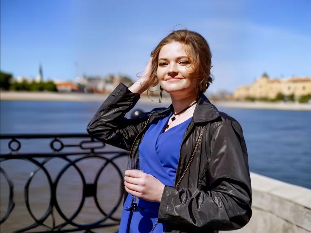 ElenaXRus heeft lichtbruin haar, draagt een zwarte jas over een blauwe jurk, en glimlacht terwijl ze met één hand in haar haar zit. Ze staat bij een zwart ijzeren hek, met een blauwe lucht op de achtergrond..