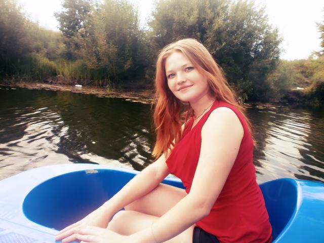 ElenaXRus met rood haar, gekleed in een rood shirt, zittend in een blauwe kajak, lachend naar de camera, met haar handen op de rand van de kajak, en een reflectie in het water..