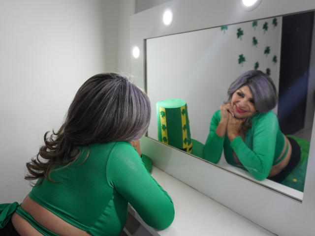 CelesteLincon with gray hair, green top, and black panties, leaning on a mirror, smiling, reflecting her ass, in a room with green decor and white walls.