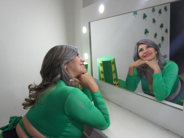CelesteLincon with gray hair, green top, and red lipstick, smiling at her reflection in a mirror, leaning on a white counter, hand on chin, green and yellow decor.