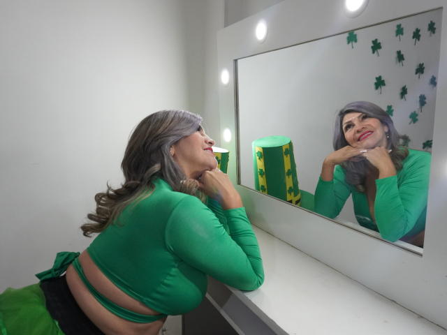 CelesteLincon with green top, curly gray hair, and red lipstick, leaning on vanity, looking at mirror, hands under chin, green and yellow box in background, smiling.